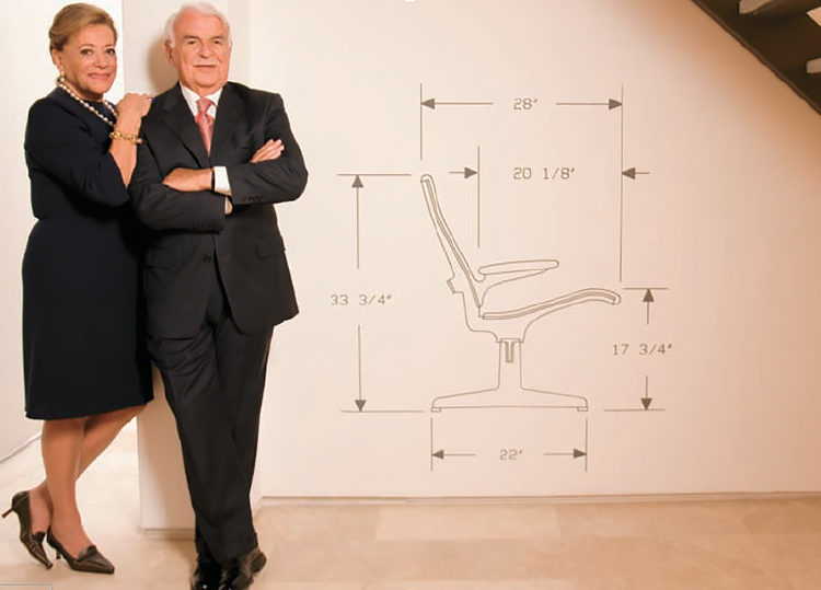 John Daniels, in a dark suit, is standing with his arms crossed against a wall next to an illustration of a chair labelled with its dimensions. Myrna stands beside him, in a black dress and heels, with one hand on his shoulder.