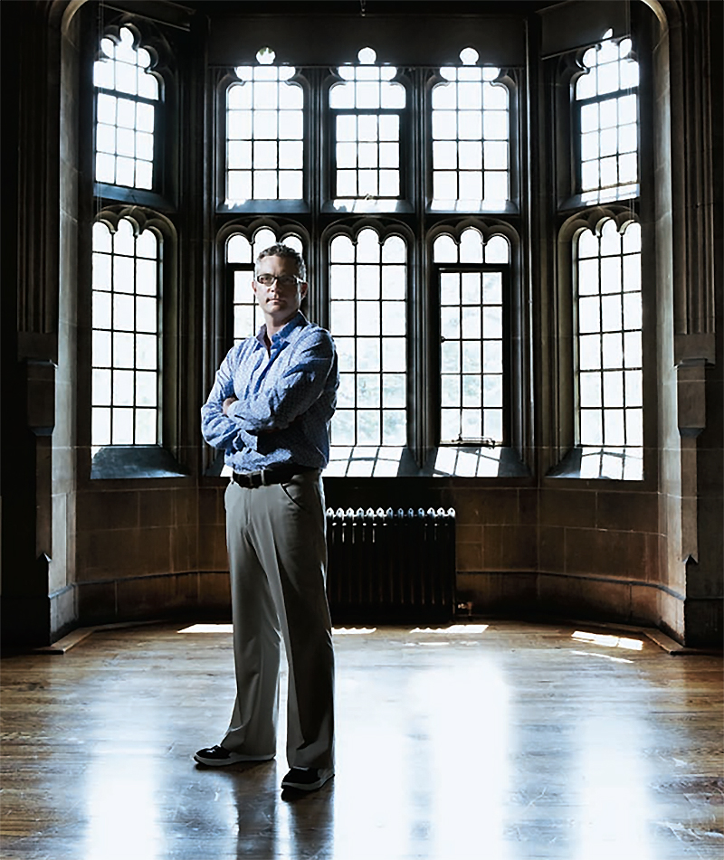 Andrew Pyper, with arms crossed over his chest, is standing in front of five columns of windows set in an alcove inside Hart House.