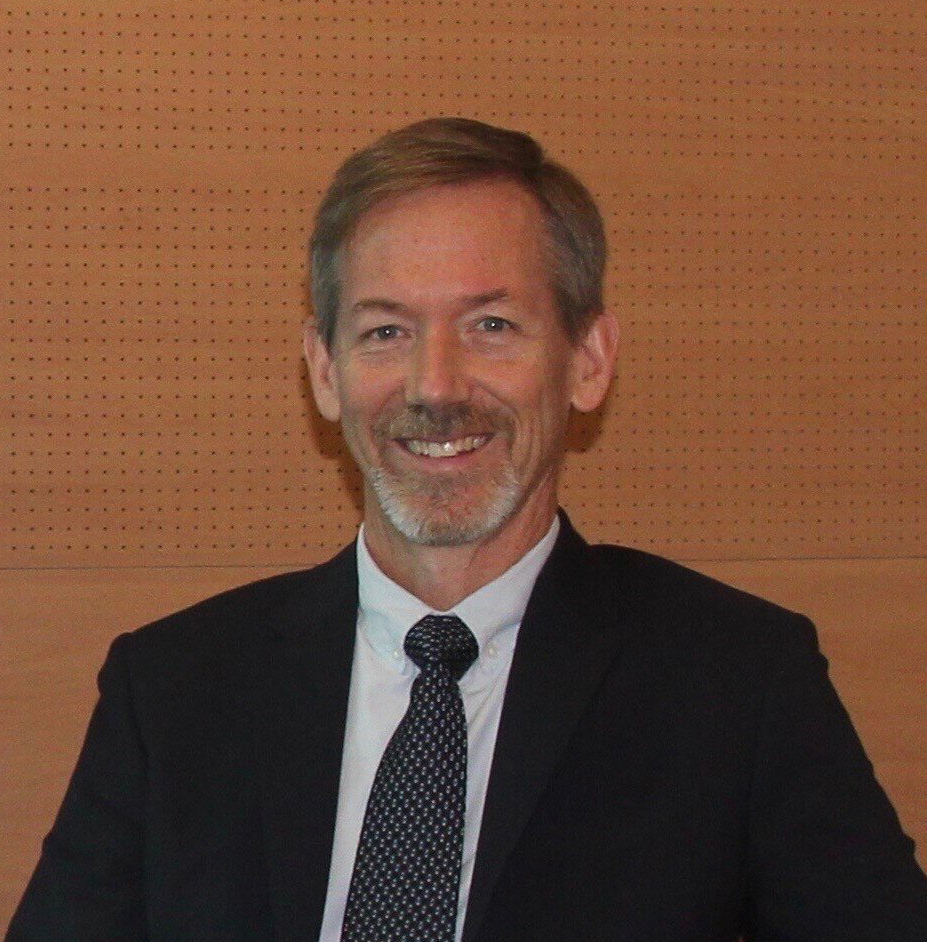 Close-up of Geoff Ibbotson in a black suit, white collared shirt and patterned tie