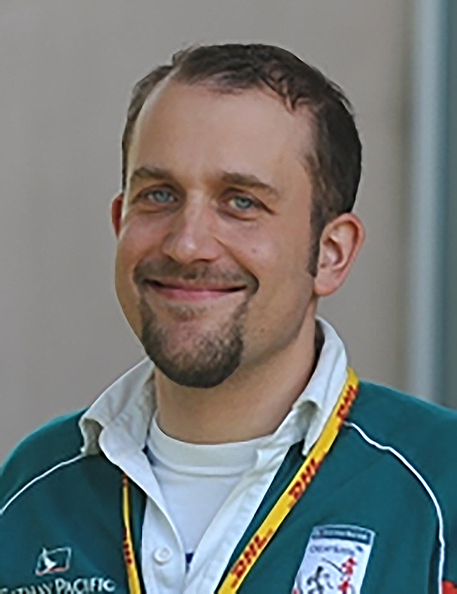 Headshot of Mark Slade wearing a green sports cardigan with logos along the front, over a white shirt