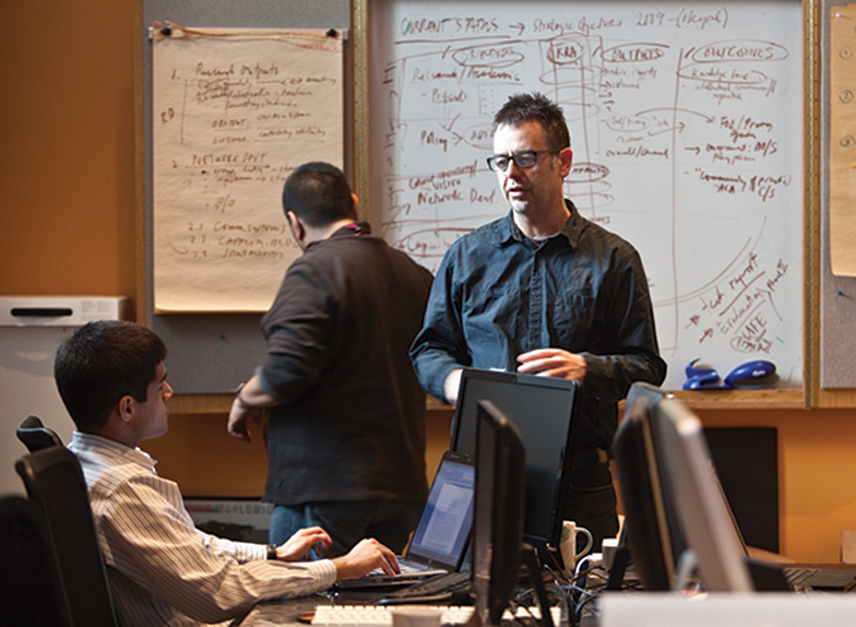 Professor Ron Deibert (centre) is speaking with a colleague, who is sitting in front of a laptop in the Citizen Lab. Another colleague is looking at notes on the wall behind him.