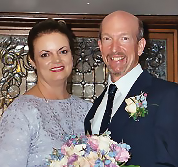 Lee Campbell, wearing a lavender dress, and Peter Forsythe in a navy blue suit and tie, with a flower decoration pinned to his lapel, are standing in front of a stained glass window, with a bouquet of mixed flowers held between them.