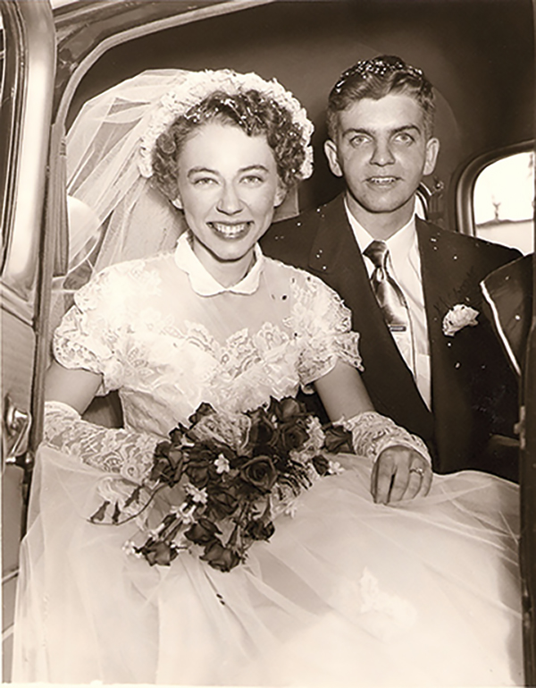 A black and white photo of Jane Brown in a white wedding gown and veil, holding a bouquet of roses, and Roger Brown, wearing a dark suit and satin tie. They are sitting in a car with the door open, smiling at the camera.