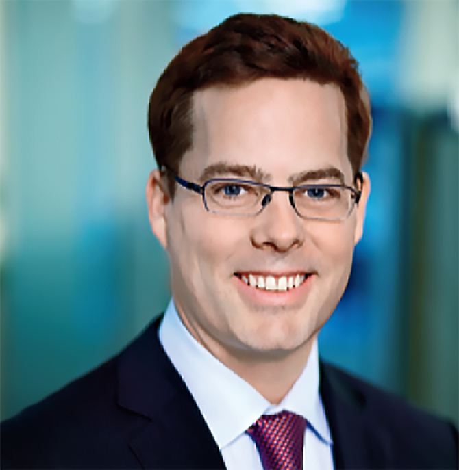 Headshot of Matt Chapman in blue framed glasses, a black suit and a purple checkered tie.