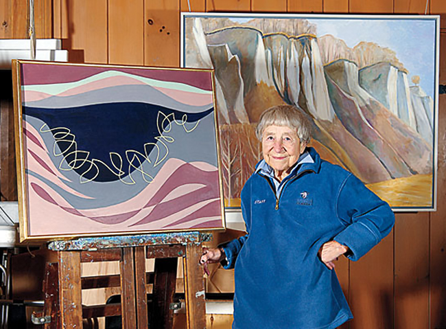 Doris McCarthy in a blue pullover, standing next to one of her abstract oil paintings on an easel. Another oil painting of cliffs hang on the wall behind her.