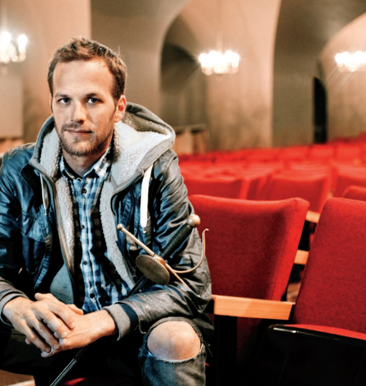 Close up of Jeremy Hutton in a leather jacket and plaid shirt, sitting on a chair in Hart theatre with a sword leaned against the inside of one arm