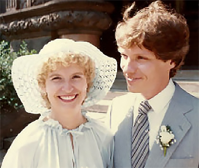 Hana Nute (on the right), wearing a white dress, large-rimmed hat and a wide smile, standing close next to Donald Nute, wearing a light grey suit, diagonally striped tie and a flower decoration pinned to his lapel