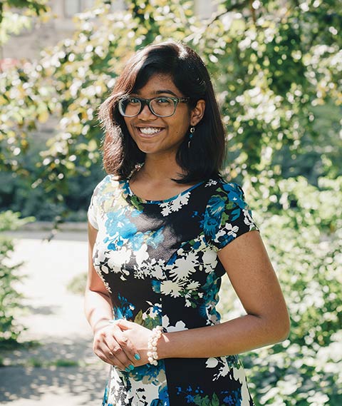 Deborah Solomon, wearing a flower-patterned dress, standing with tree leaves out of focus in the background