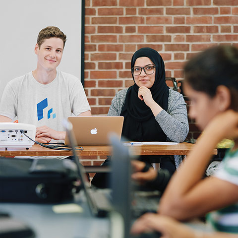 Will Ginsberg and Afifa Saleem sitting at a desk in front of an open laptop, a student out of focus in the foreground