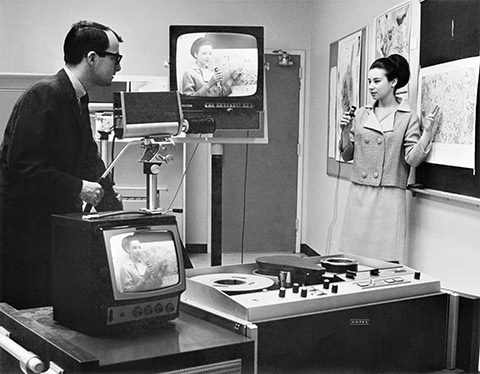 Photo of a student speaking into a hand-held microphone and pointing to a chart. A teacher is recording the student, and the image is displayed on two television screens.