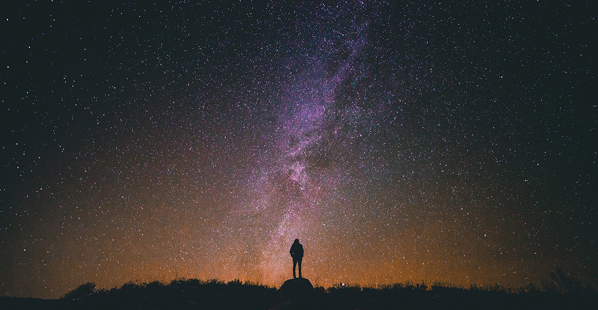 Small silhouette of a figure standing on a rock looking up at the night sky