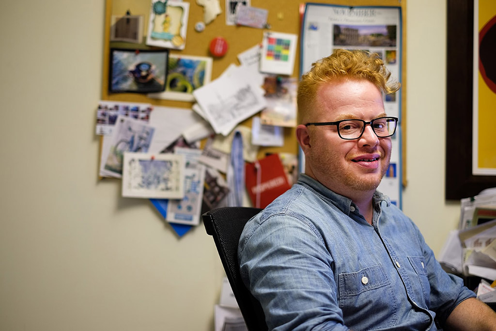 Close up photo of David Roberts sitting in a chair on the right side of the frame, facing right, with a bulletin board covered with cards and papers in the background