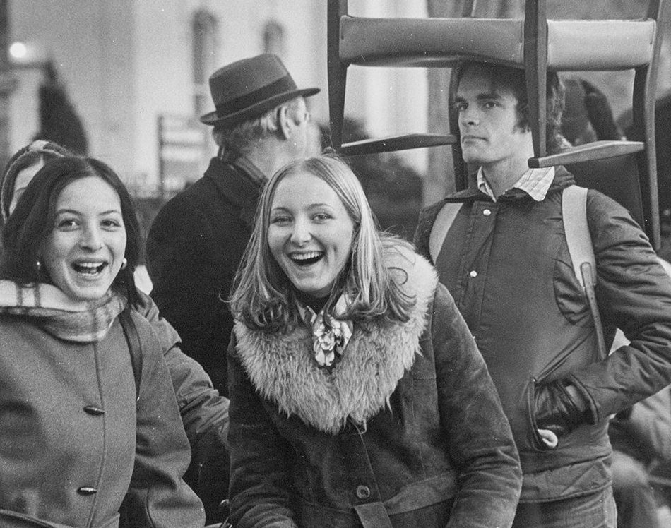 Black and white photo of students in the parade. Two young women in front are smiling and a young man standing behind them is carrying a chair over his head.