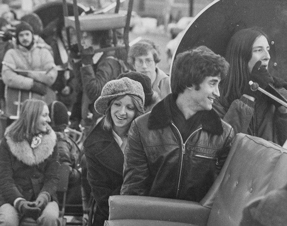 Black and white photo of students standing in line in the parade, carrying chairs and a couch