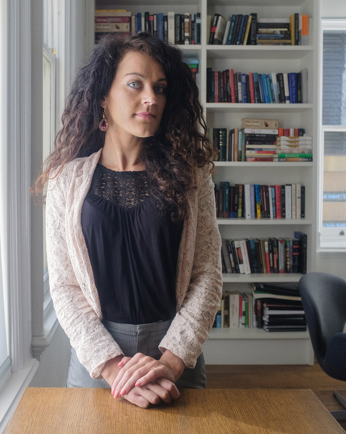 Photo of Petra Molnar standing next to a window in front of a desk, with a bookshelf in the background