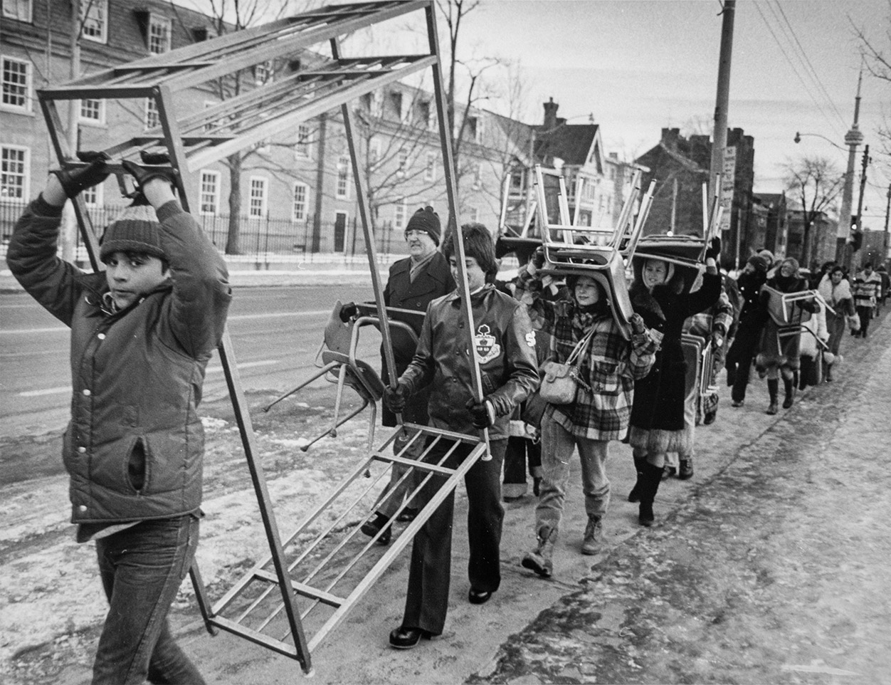 Black and white photo of students marching in line and carrying chairs. Two students upfront are carrying a metal rack.