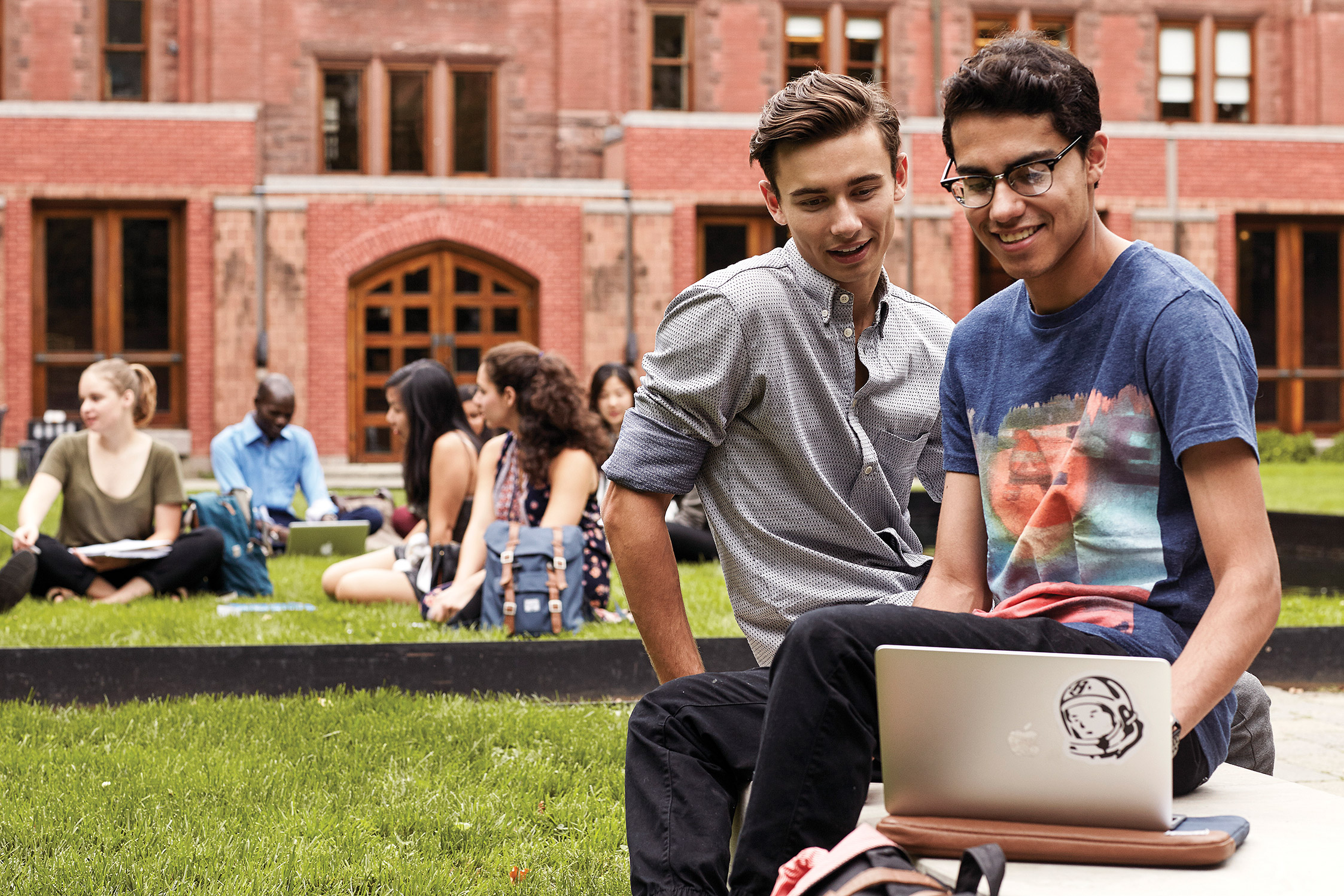 Students Quinn Underwood and Swarochish Goswami are working on a laptop while seated on a bench outdoors. Other students are sitting on grass in the background.