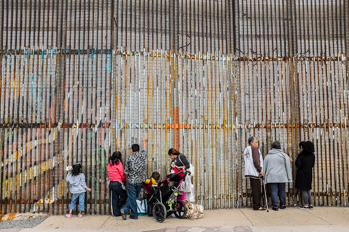 In the foreground, a family of five, with two young girls and a toddler in a stroller, stand facing the border gates in the background. An elderly couple and another adult stand a few feet away also facing the gates.