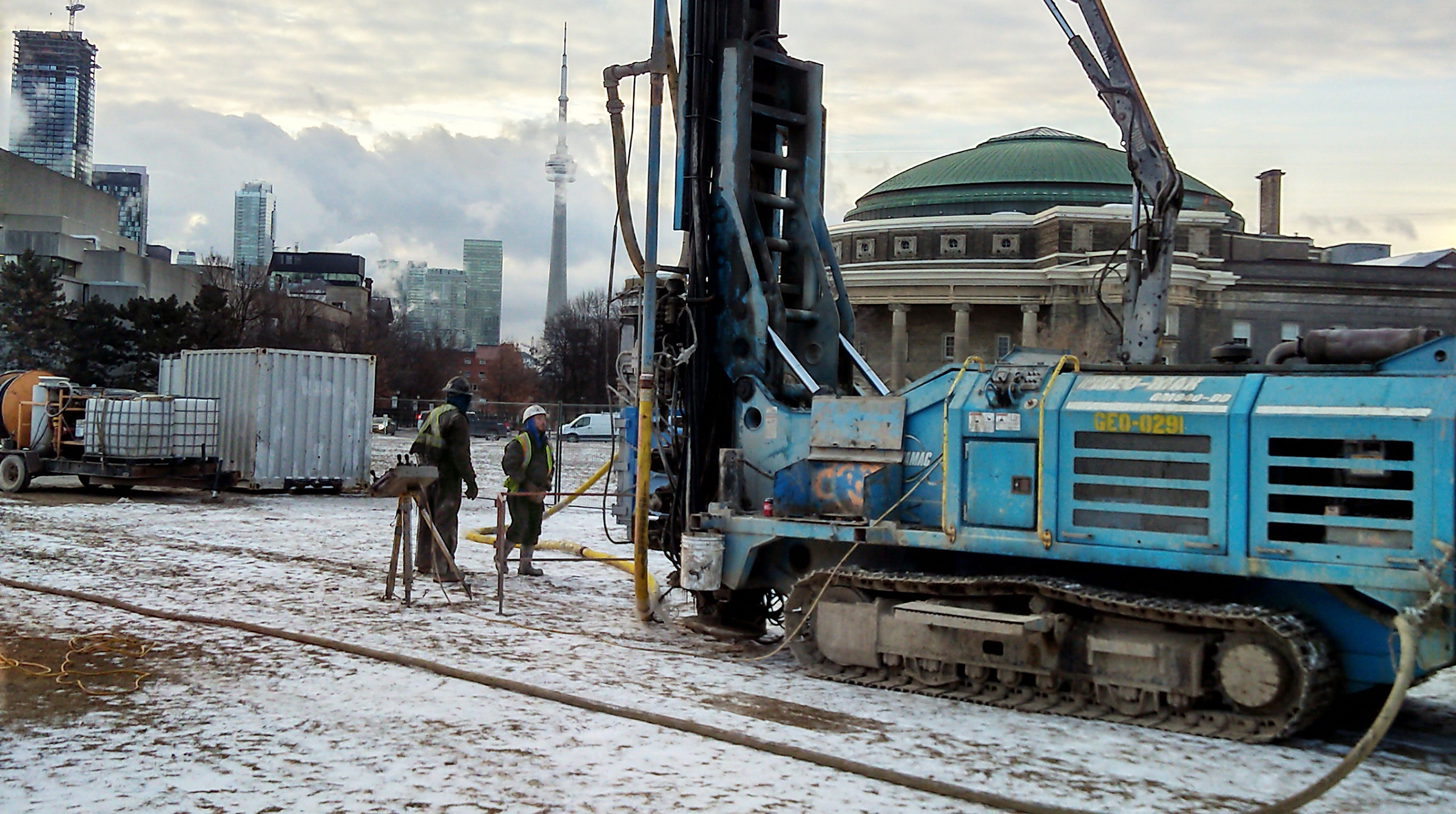 Two workers standing in front of a tank-like machine drilling a hole into the ground of the front campus field