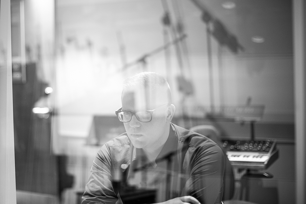 Black and white photo of Eliot Britton sitting in a music studio behind glass that is reflecting rod-like shapes