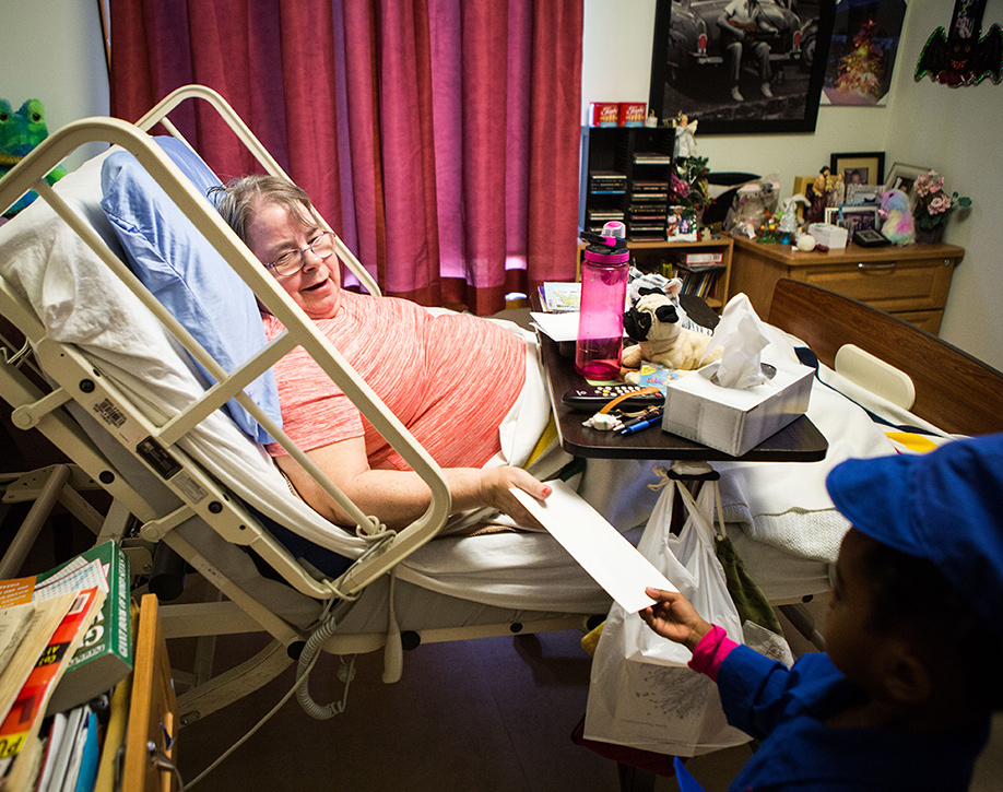 A young child in a blue uniform handing a letter to an elderly woman, who is lying semi-reclined in a hospital bed with a tray above her lap