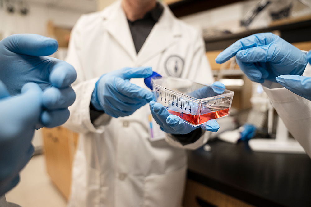 Close up of the blue gloved-hands of three scientists, one of whom is holding a transparent container with clear red liquid on its side