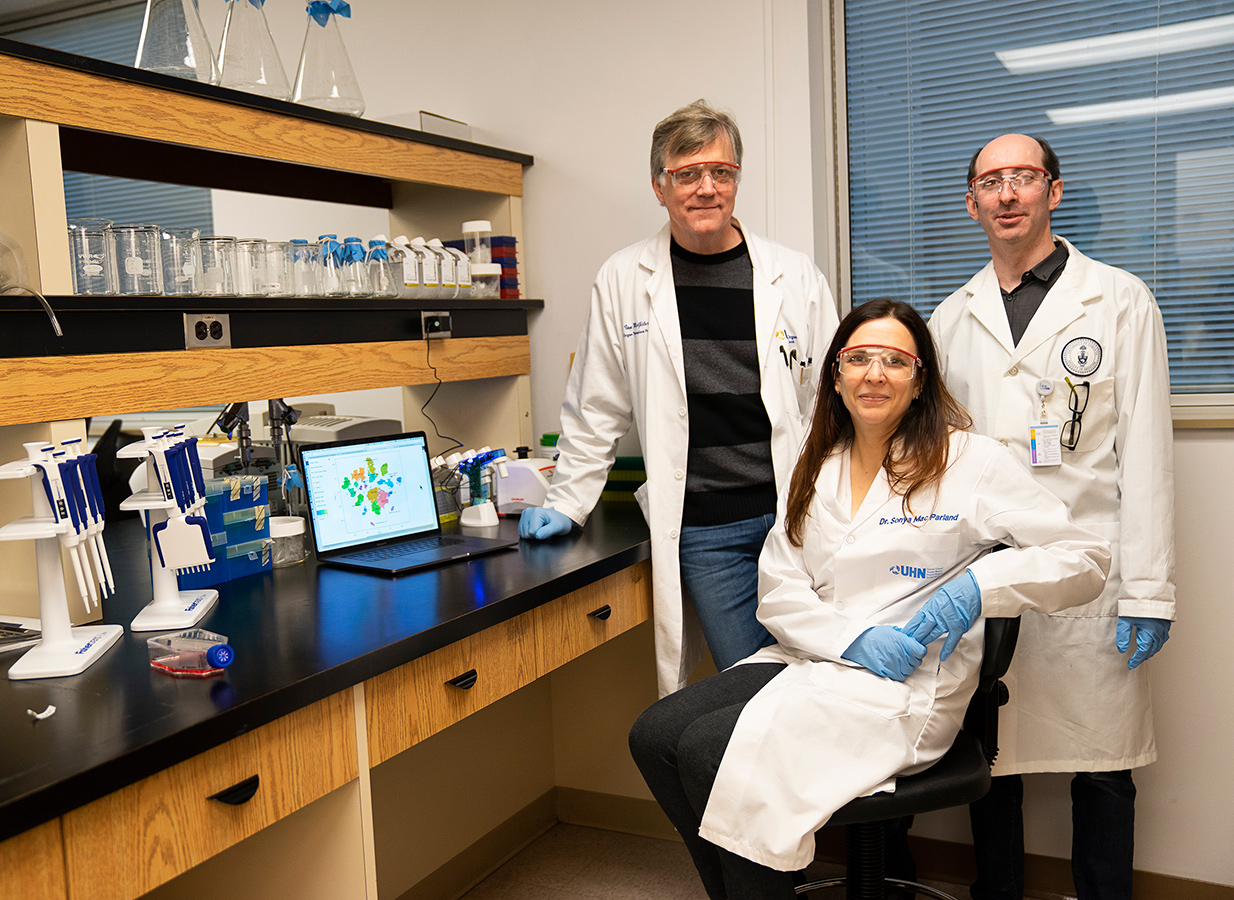 Ian McGilvray and Gary Bader standing off-centre, with Sonya MacParland sitting in a chair in front of them, all wearing lab coats and safety glasses. McGilvray is in the centre of the photo, leaning against a table with glass beakers and an open laptop.