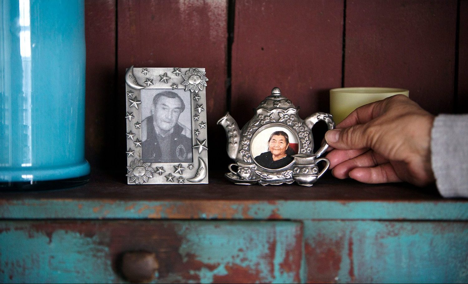 Close up shot of a black and white photo of Harold Wastasecoot in a metal frame containing stars, moons and suns, and a colour photo of Maria Wastasecoot in a metal frame in the shape of a tea pot. The photos are resting on a shelf next to a blue ceramic cylindrical container on the left side, and a hand is holding the handle of the teapot on the right side.