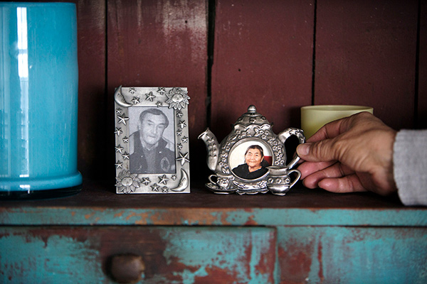 Close up shot of a black and white photo of Harold Wastasecoot in a metal frame containing stars, moons and suns, and a colour photo of Maria Wastasecoot in a metal frame in the shape of a tea pot. The photos are resting on a shelf next to a blue ceramic cylindrical container on the left side, and a hand is holding the handle of the teapot on the right side.