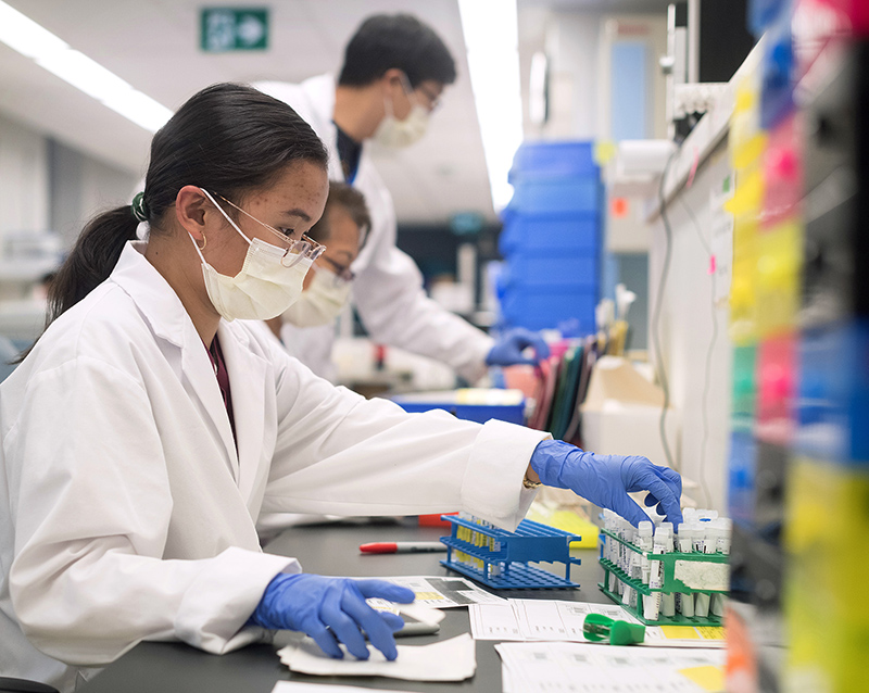 U of T researchers in lab coats and masks working at desks with test tubes and note papers