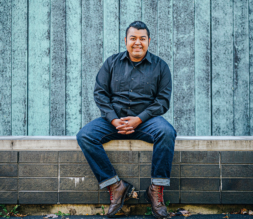 Prof. Jerry Flores smiling and sitting on a bench outside with the blue wooden walls of a building behind him