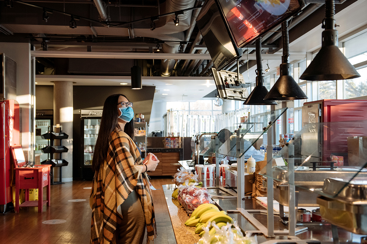 Shirley Liu looking up at the menu screen in the cafeteria in front of the counter, where bananas and individually packaged apples are laid out