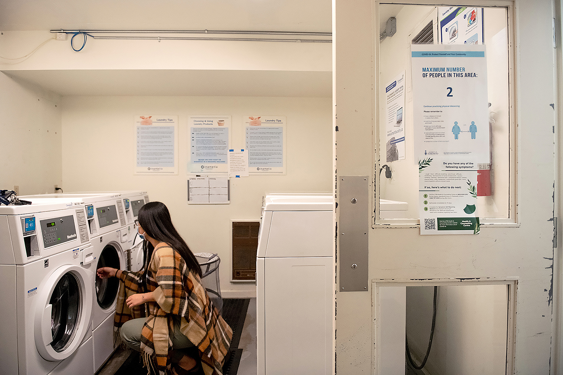 Shirley Liu crouched in front of washing machine in a laundry room with a sign on the door indicating a maximum of two people allowed in the area