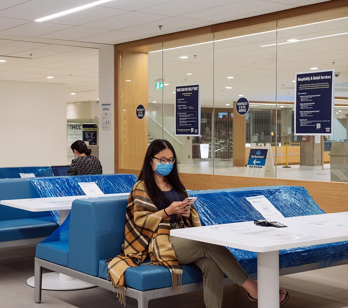 Shirley Liu in a student lounge, looking at her phone while sitting on a blue bench half-covered with plastic wrap