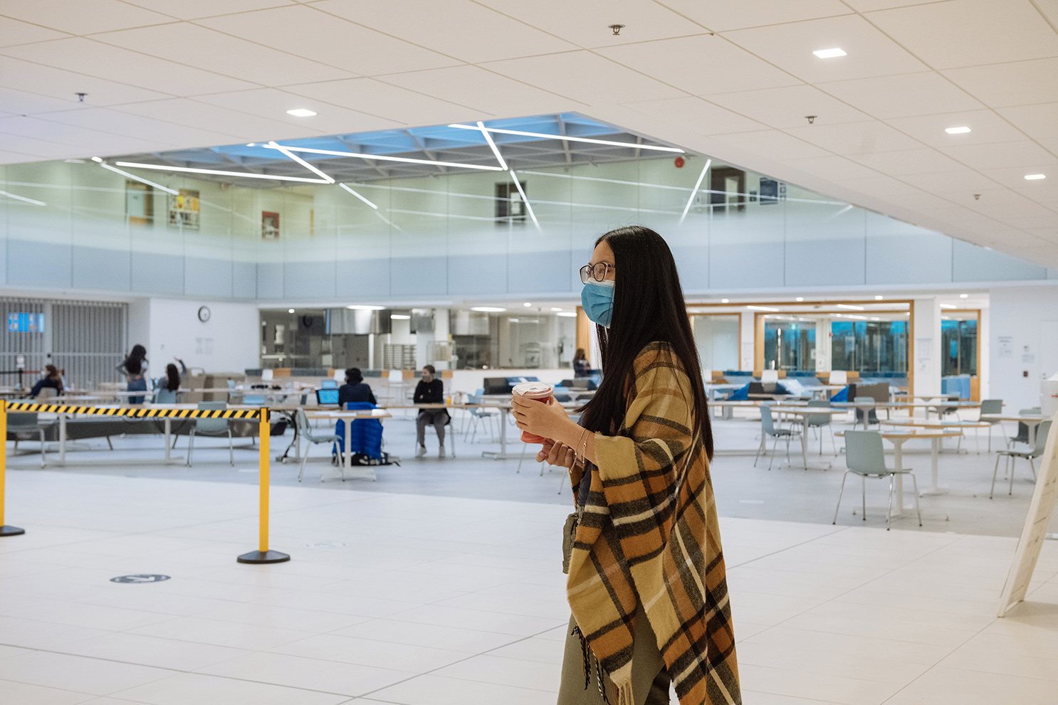 Shirley Liu in a plaid shawl and face mask carrying a coffee cup and walking by a student lounge within the Davis Building