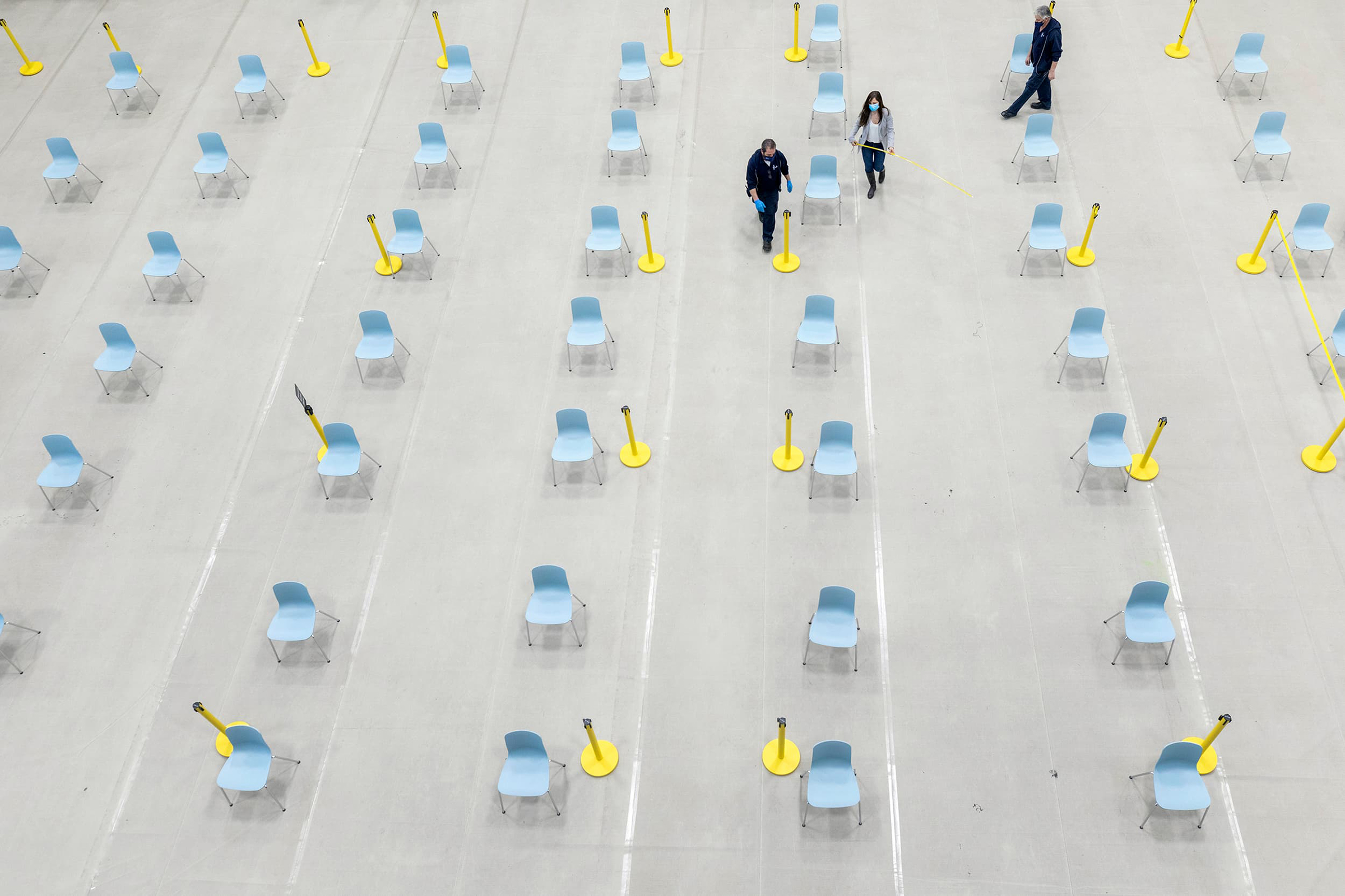 Rows of empty chairs at the vaccination site and three staff members wearing masks near the top of the photo, one holding a tape measure