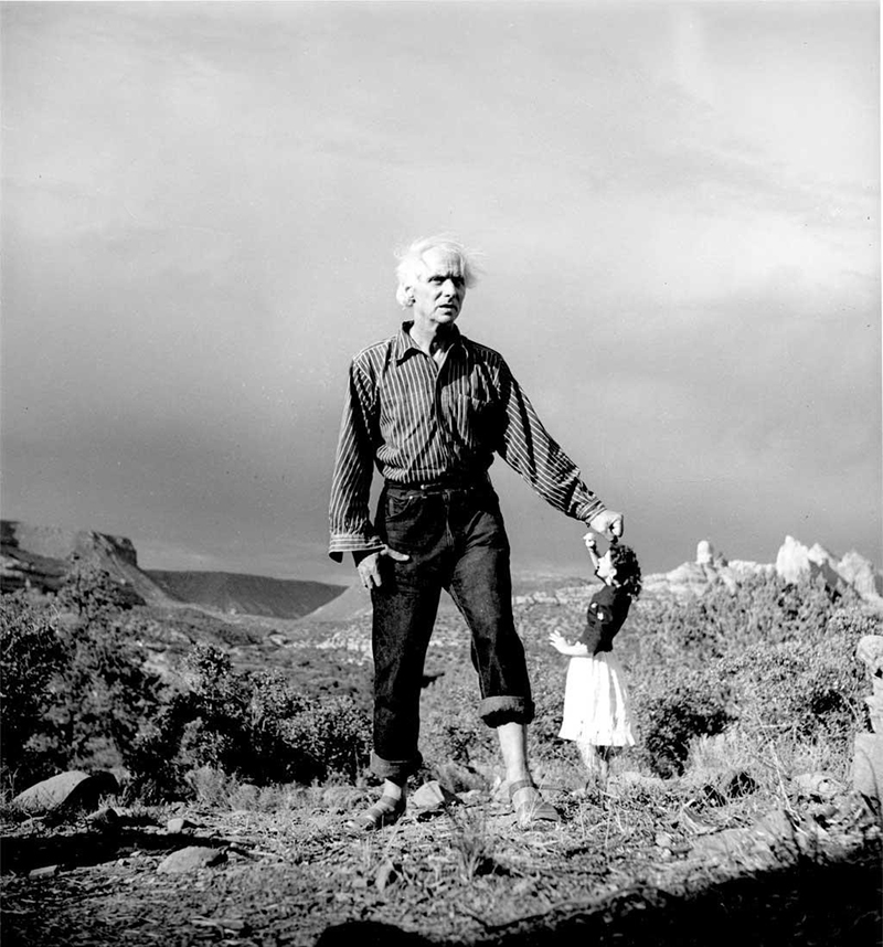 Black and white photo of a giant-sized man standing on a grassy landscape beside a miniature-sized woman looking up at him