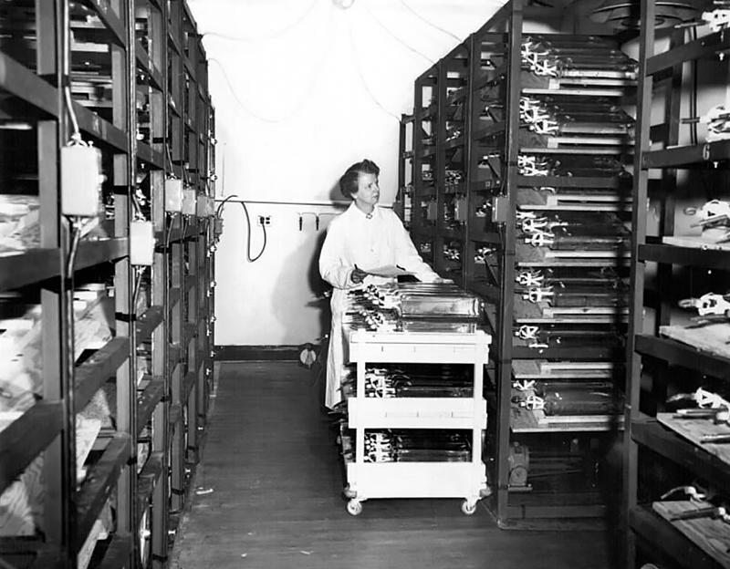 Black and white photo of Leone Farrell in a lab coat looking at items on a storage shelf