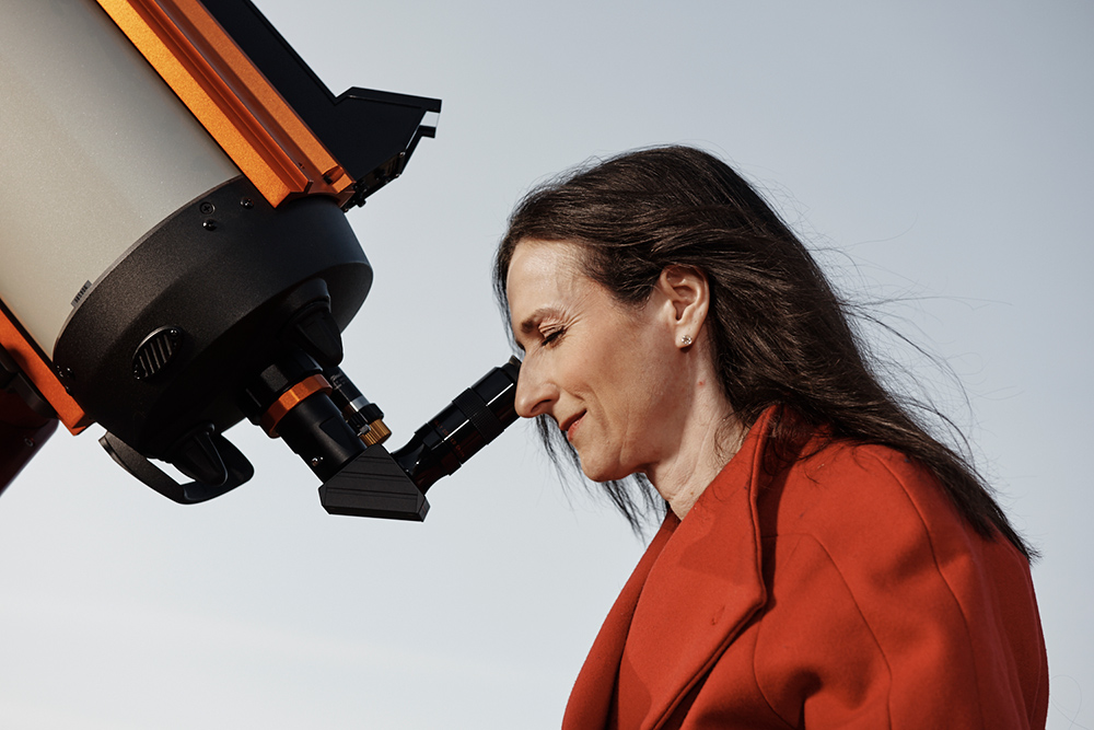 Sara Seager, who has dark hair and is wearing a red coat, is in profile, peering through the eyepiece of a telescope