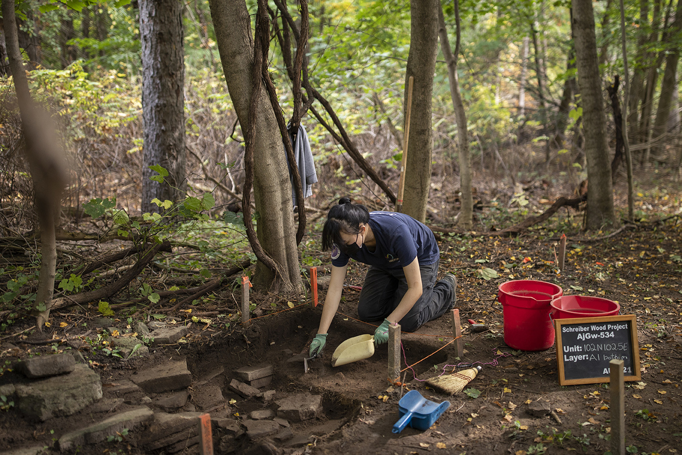 U of T Mississauga student digging soil with a hand shovel in a rectangular section of the forest marked by wooden sticks