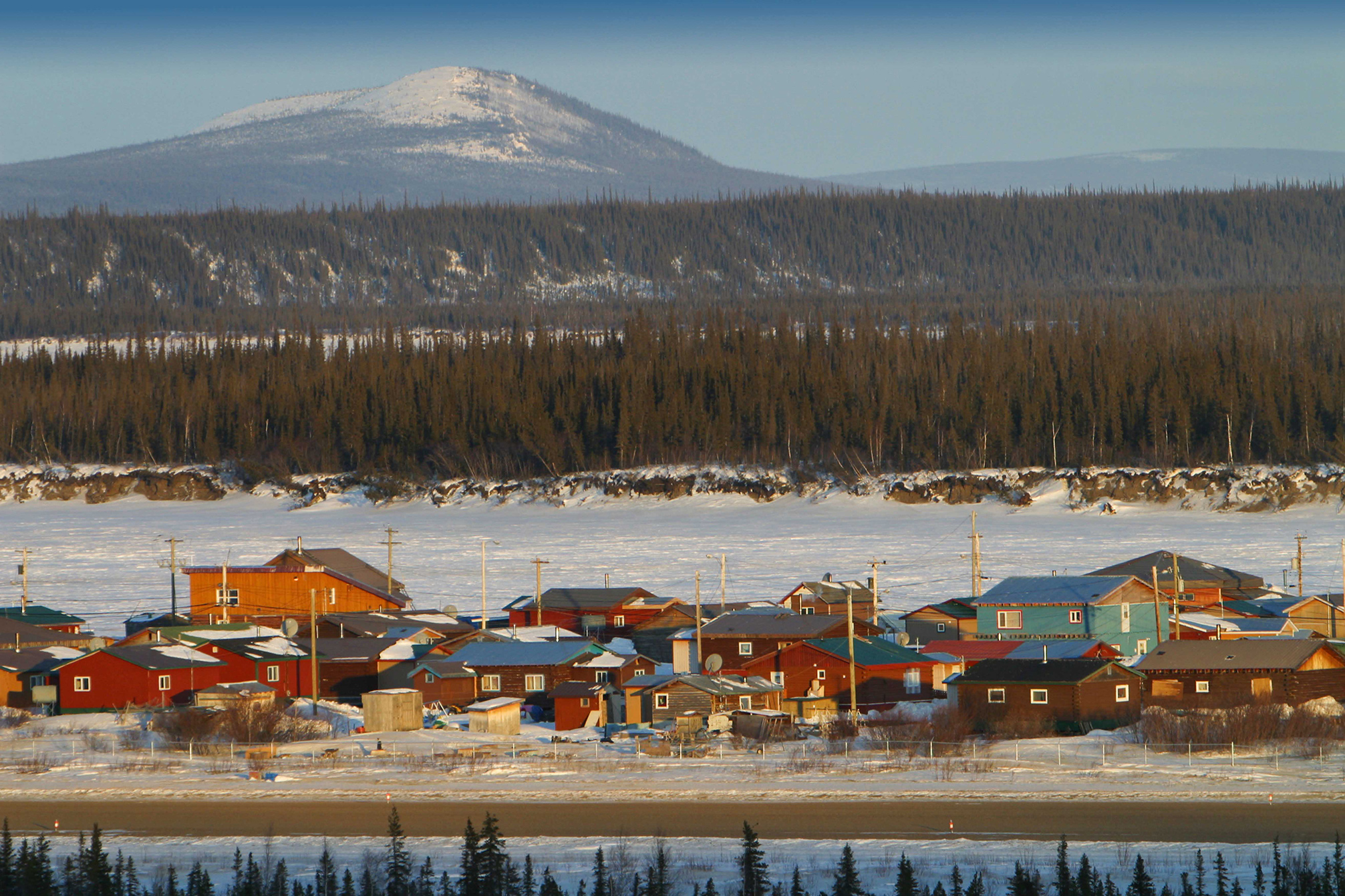 Houses in Old Crow flanked by a road and a field of snow on the other side, surrounded forest and a mountain in the distance