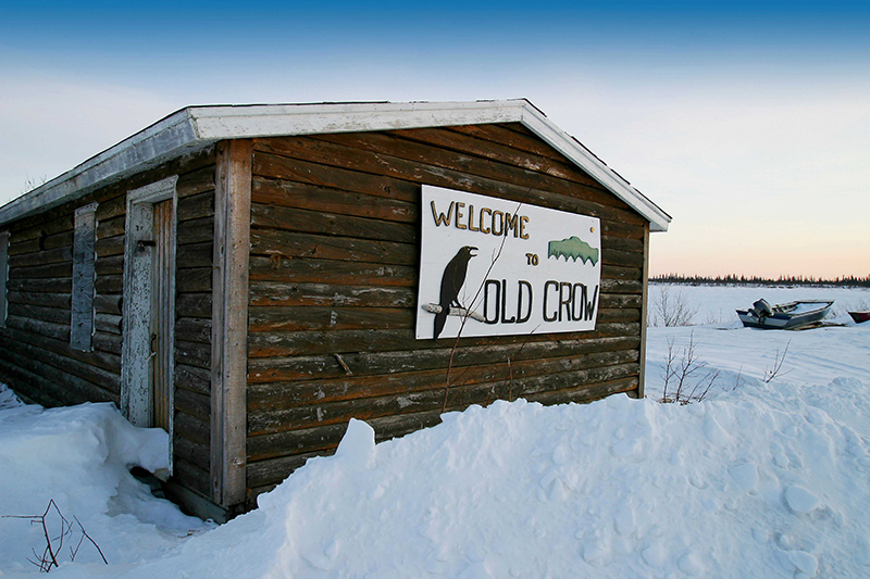 "Welcome to Old Crow" sign on the side of a log shed surrounded by snow