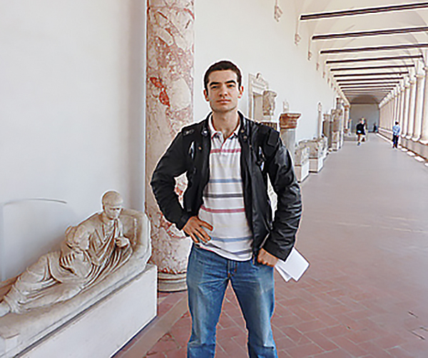 Ilya Sutskever in a black jacket and blue jeans standing beside a statue of a reclining robed figure inside a museum.