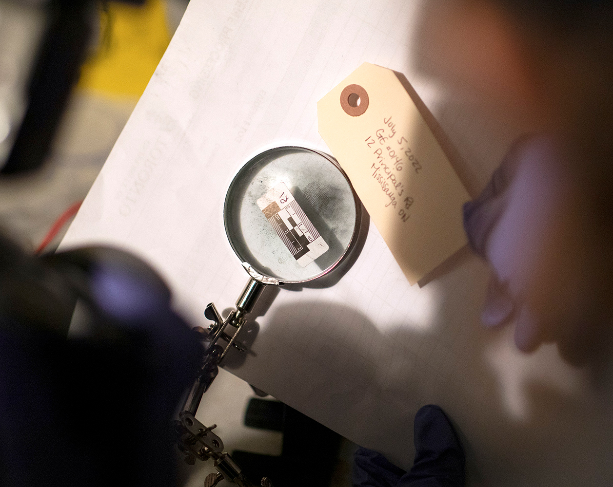 A magnifying glass with a ruler in the centre is placed on top of dark thumbprints on white paper, while a blue-gloved hand holds a paper tag next to it, handwritten with a date, GE number and location