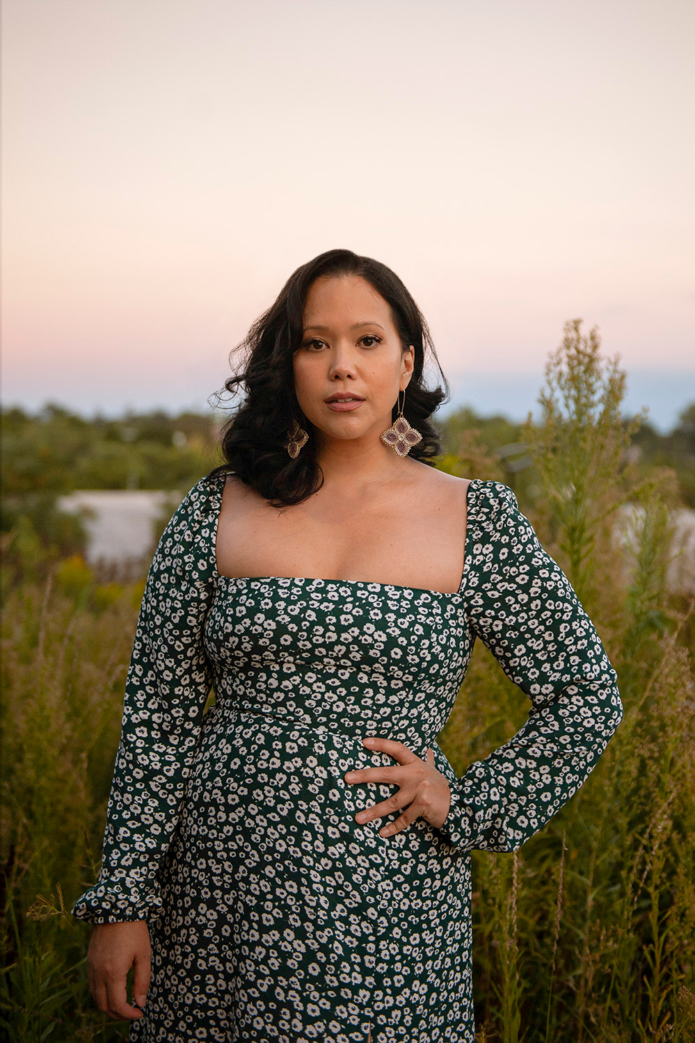 Prof. Kristen Bos wearing a long-sleeved, black and white flower patterned dress and large purple clover-shaped earrings, standing in front of a field of tall shrubs, with one hand on her waist