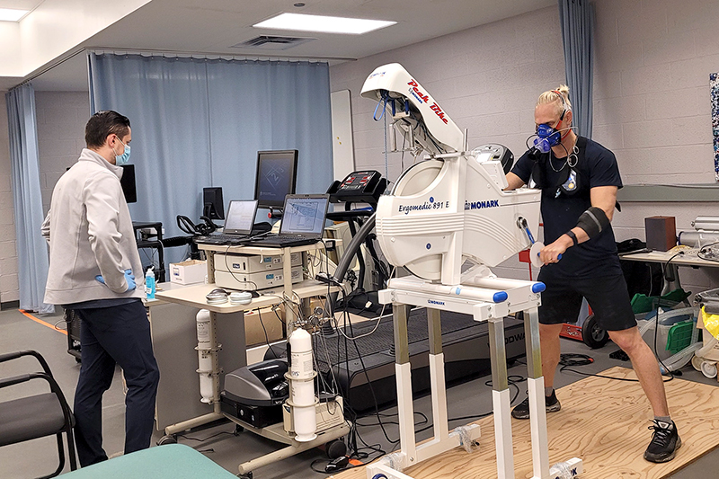 A researcher in a blue face mask and gloves observes data on laptops as a research subject wearing an oxygen mask pedals a machine with a wheel with his hands