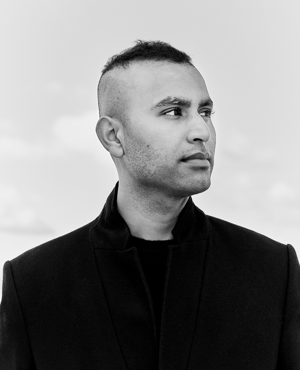 Black and white headshot of Jiavet Ealom in a black overcoat, looking off to the right