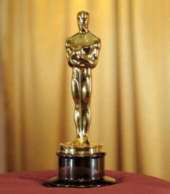 Close up of an Oscar award trophy resting on top of a red pillow, with gold-coloured curtains in the background