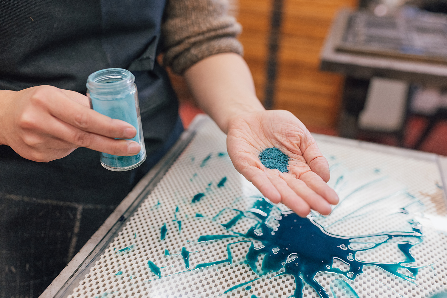 A hand holds out blue-green crystals over a table top with a lot of crystals on it. Another hand holds a small bottle of the crystals.