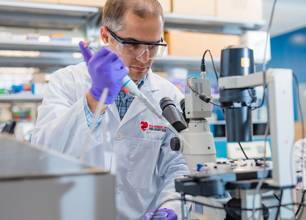 A researcher wearing a white lab coat, blue disposable gloves and protective eyewear, working with a machine in a lab at the Ted Rogers Centre for Heart Research and holding a pipette